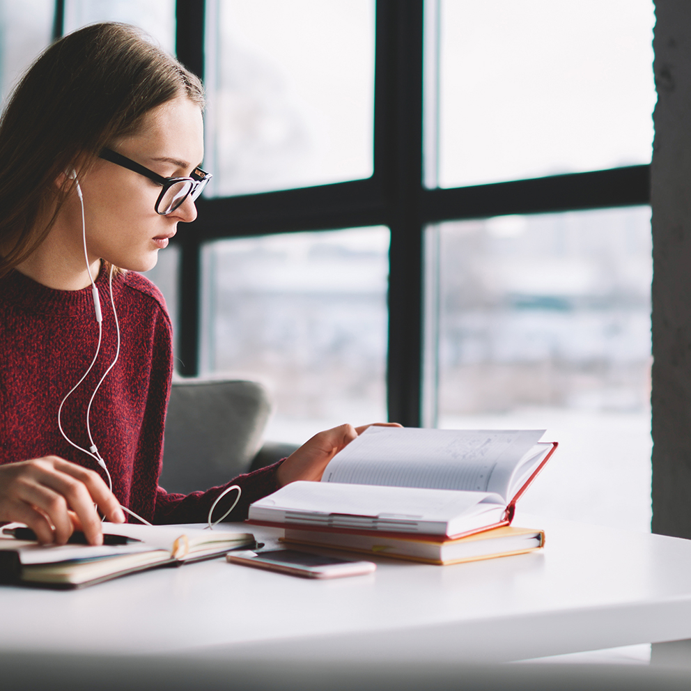 Attractive hipster girl learning at university library while enjoying favourite music via headphones, young female student reading book and preparing for exam with textbook enjoying playlist