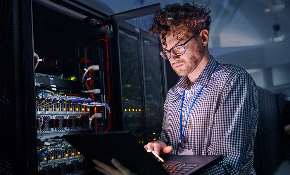 Focused male IT technician working at laptop in dark server room