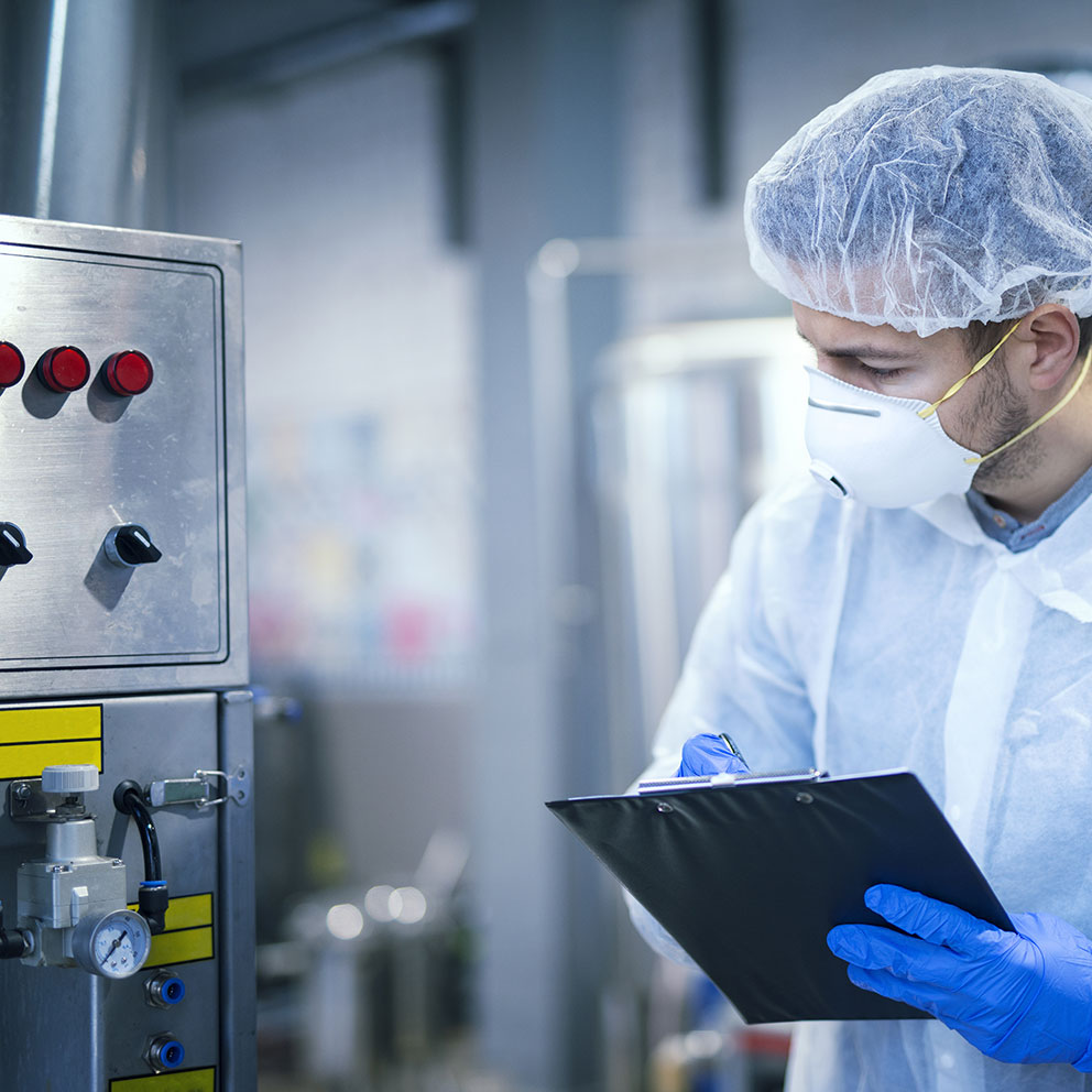 Technologist expert in protective uniform with hairnet and mask taking parameters from industrial machine in food production plant.