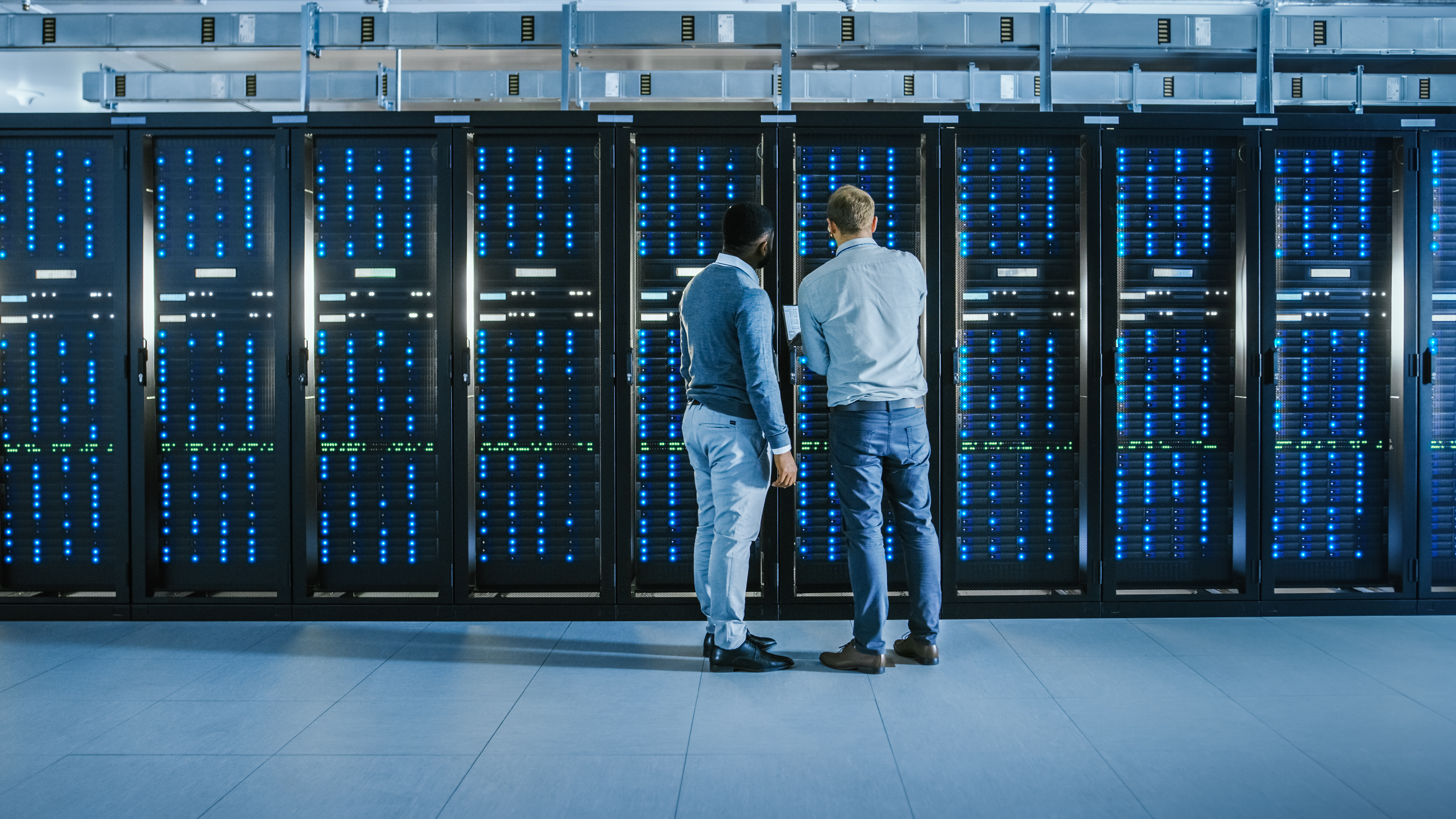 Bearded IT Technician in Glasses with a Laptop Computer and Black Male Engineer Colleague are Talking in Data Center while Working Next to Server Racks. Running Diagnostics or Doing Maintenance Work. 